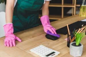 lower half of a body wearing forest green apron and pink cleaning gloves wiping a computer mouse on a wooden desk workstation